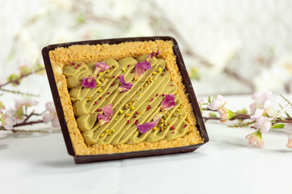 Mazurek z kremem pistacjowym – kruchy spód z prażynką i białą czekoladą, pokryty warstwą malinowego musu oraz kremem pistacjowym z białą czekoladą. Dekoracja z jadalnych płatków róż i posiekanych pistacji. Domowy wypiek cukierni Jacek Placek, przygotowany bez dodatków chemicznych z naturalnych składników, idealny na Wielkanoc, catering i do kawiarni.